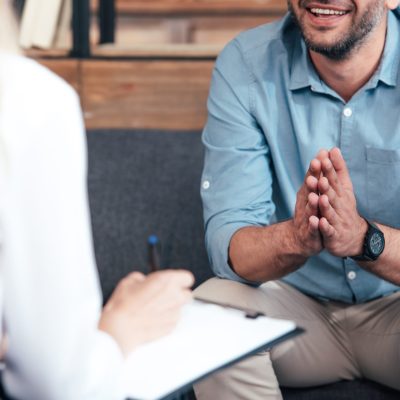 cropped image of female counselor writing in clipboard and smiling male holding hand palms together during therapy session in office